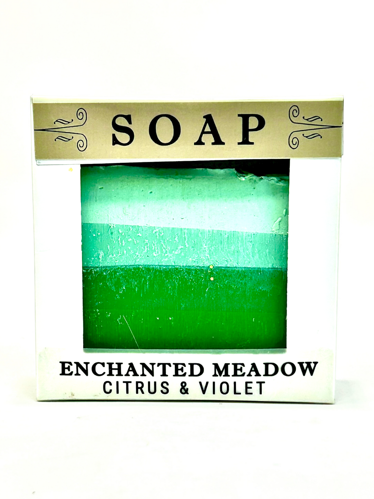 A boxed soap bar labeled 'SOAP' and 'ENCHANTED MEADOW CITRUS & VIOLET,' showing a green and aqua striped soap inside a white package with a gold banner.